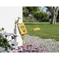 Ein gelber Gartenbewässerungstimer ist an einen Wasserhahn angeschlossen, während im Hintergrund ein Rasensprenger in einem Garten läuft.